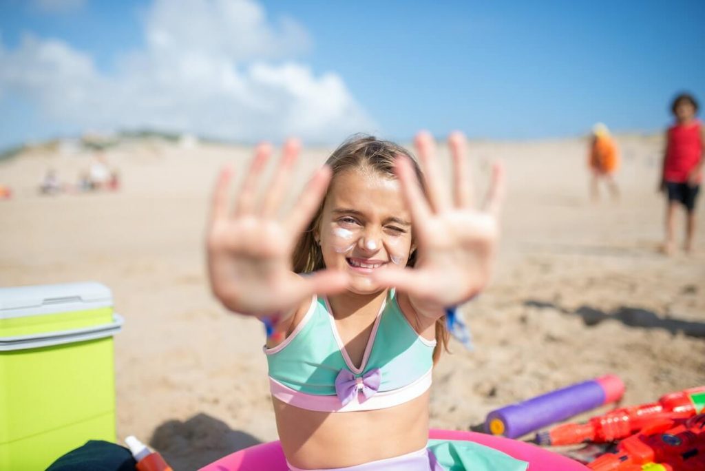 Devojčica sa kremom na licu sedi na plaži i pokazuje ruke kameri