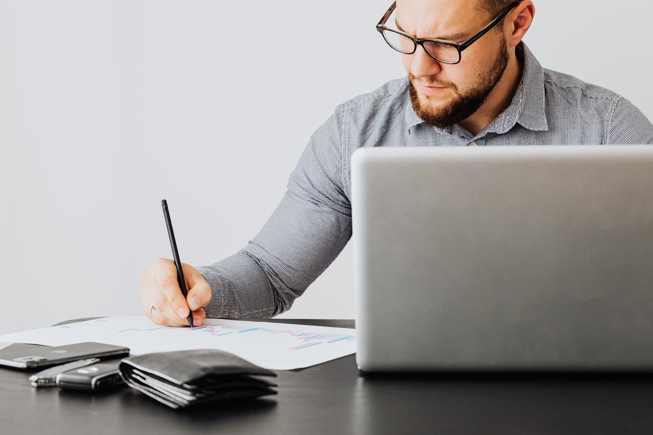 Čovek sedi za laptopom sa papirima i olovkom.